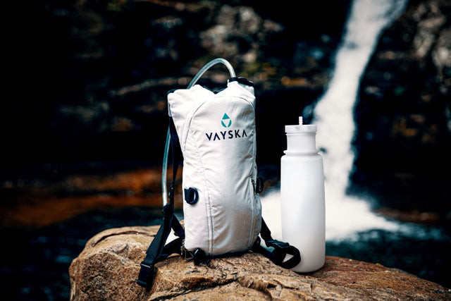 White hydration backpack with the bottle beside the pack against a waterfall backdrop.