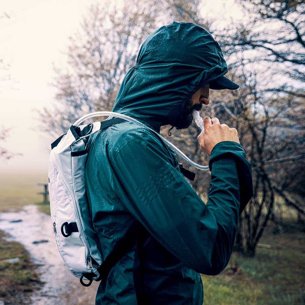 A young outdoor enthusiast takes a drink from his white Vayska hydration backpack during a rain storm.