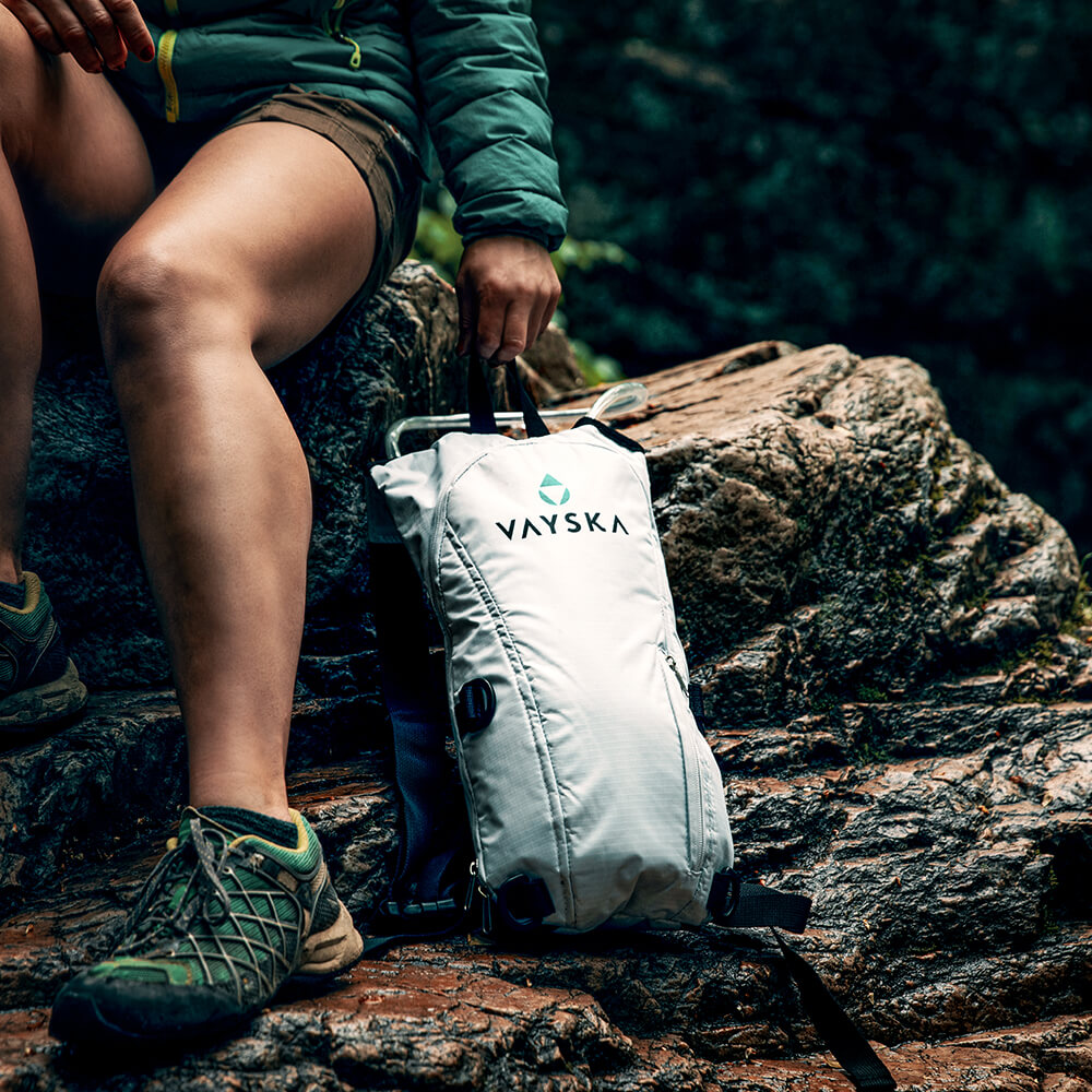 A young hiker rests on a rock carrying a white Vayska hydration backpack after hard hike up a trail.